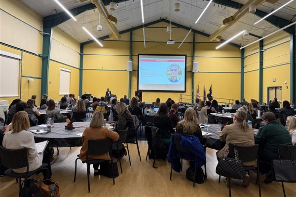 Attendees sit at round tables viewing a large screen at the end of the room.