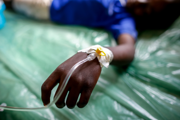 The small hand of a Black person lays on a background of a green shiny blanket with an iv tubing coming out of the top of the wrist, held in place by white medical tape.