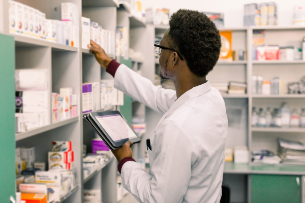 A Black pharmacy worker wearing a white coat reaches toward a bottle of medication on a shelf, while holding an electronic tablet in their other hand.